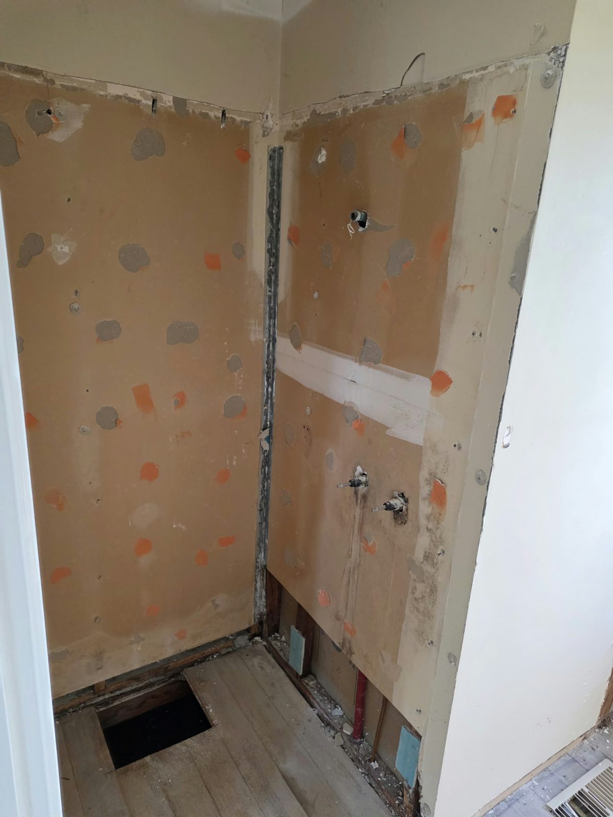 Shower zone during the Belmont bathroom strip-out — patched wall substrate, shower connections and an open floor access point
