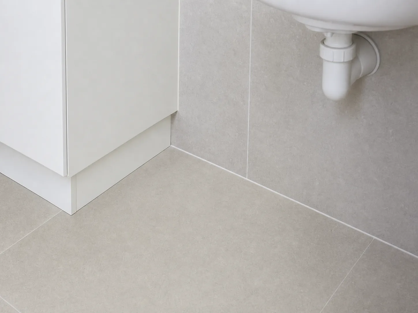 Laundry floor meeting a tiled wall with a sealed junction beside the cabinet base under the trough