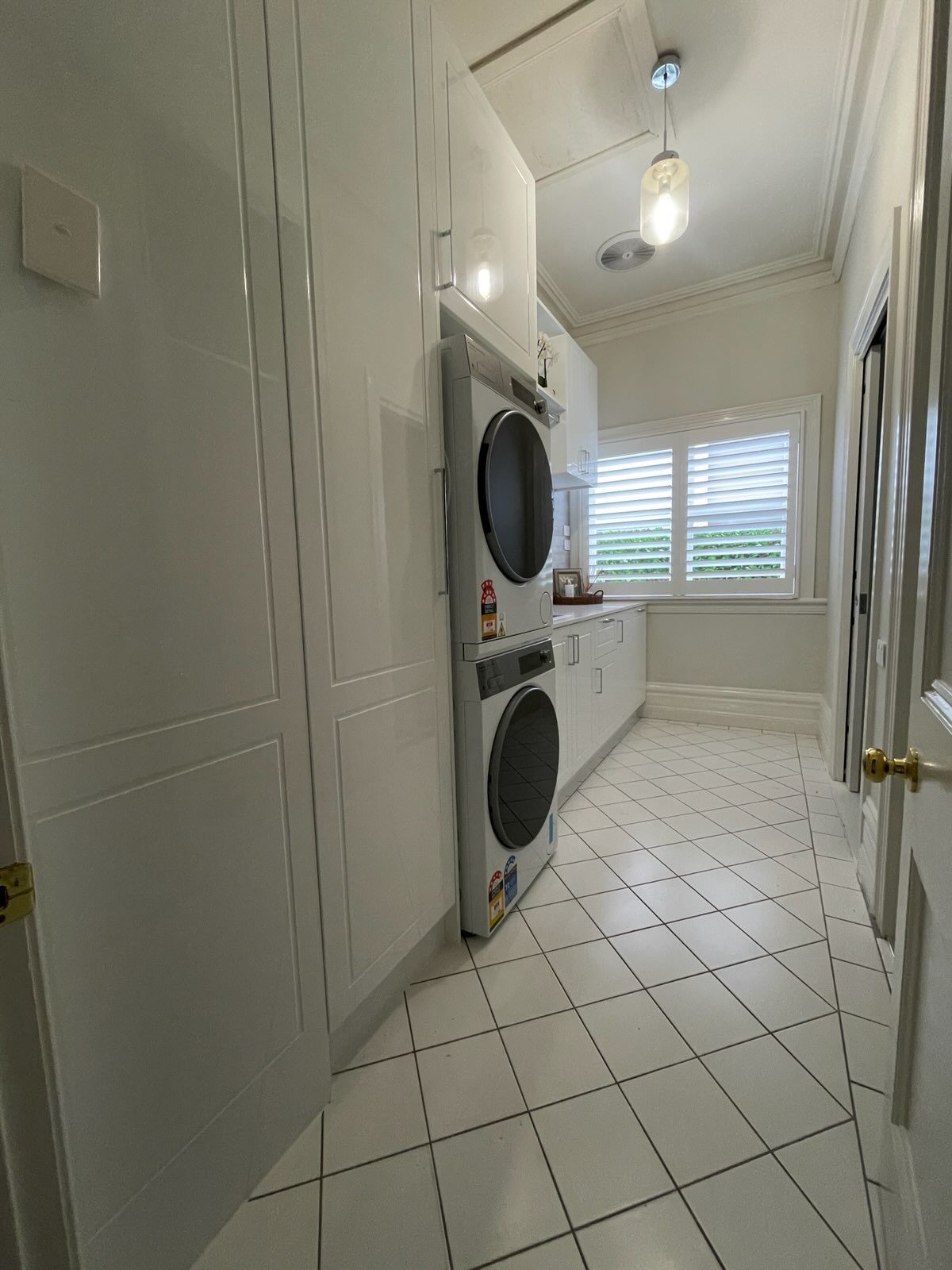 Laundry renovation by Ultimate Renovations — narrow elongated room viewed from doorway, stacked appliances at far end, white cabinetry and diagonal floor tiles