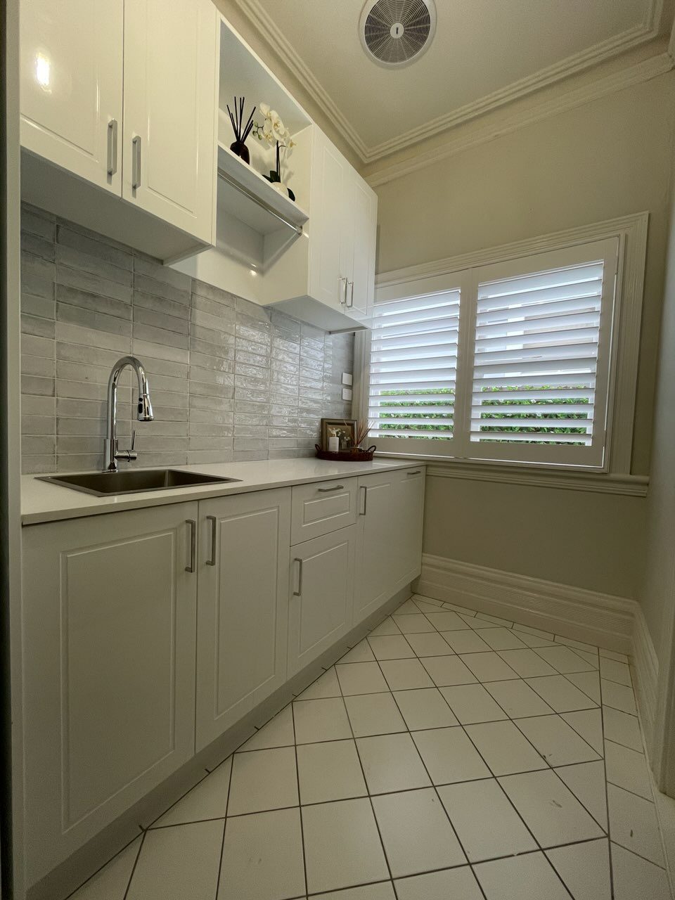 Laundry overhead cabinetry with open display shelf, stainless-steel sink, white benchtop, chrome high-arc tap and plantation shutters — Ultimate Renovations Geelong